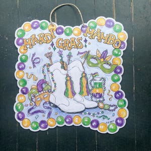 May include: A decorative Mardi Gras sign with a rope hanger. The sign features white cowboy boots, masks, and beads, with the words "Mardi Gras Mambo" in gold lettering. The border is adorned with green, purple, and gold beads.