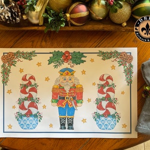 May include: A white placemat with a Christmas theme featuring a nutcracker, candy canes, and holly. The placemat has a white background with red, green, and blue accents. The nutcracker is wearing a red and blue uniform and holding a sword. The candy canes are stacked in a tower and are decorated with green leaves. The holly is green with red berries.