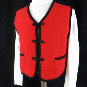 May include: A red vest with black trim and frog closures. The vest has two pockets and is worn over a white long-sleeved shirt. The vest is made of a textured material.