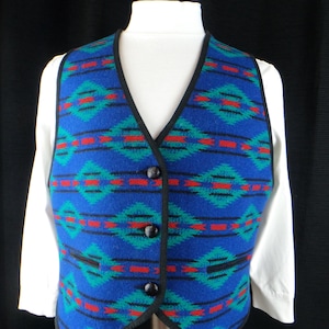 May include: A blue vest featuring a geometric pattern in teal and red with thin black horizontal lines. The garment has a V-neckline, black trim along the edges, and three black buttons down the front, worn over a plain white long-sleeved shirt.