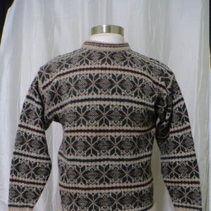 May include: A beige, long-sleeved sweater with a crew neck and a snowflake pattern in navy, burgundy, and dark brown. The sweater is displayed on a mannequin. The cuffs and hem are ribbed.