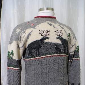 May include: A grey and cream knit sweater featuring a winter landscape with two dark deer, pine trees, and snowflakes. The garment has a red trim at the crew neckline, cuffs, and hem, with a textured cable knit pattern on the lower section.