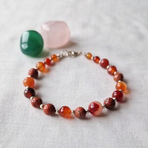 May include: A red, orange and brown beaded bracelet with a silver clasp. The bracelet is made with natural stones and is a beautiful piece of jewellery.