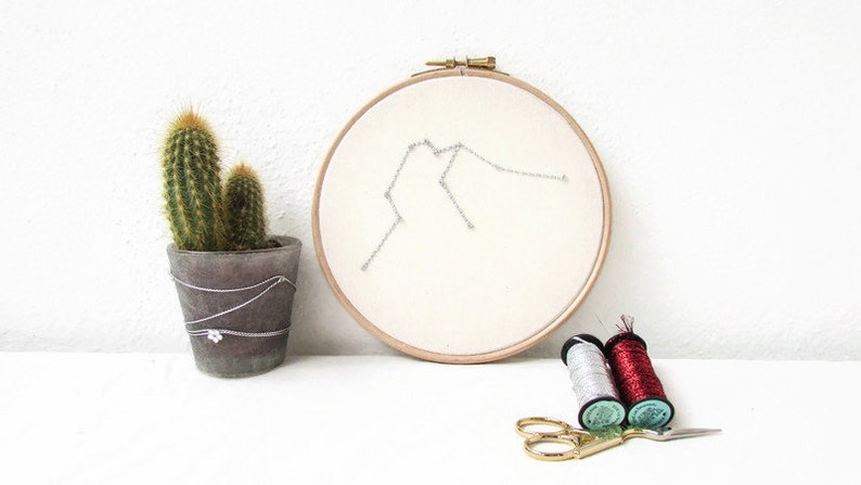 May include: A white embroidery hoop with a silver thread design of the Aquarius constellation. Two spools of thread, one red and one silver, are on a white surface with a pair of gold scissors. A small cactus plant is in a grey pot.