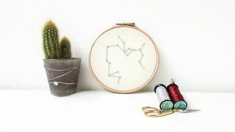 May include: A white embroidery hoop with a silver thread design of the Sagittarius constellation. Two spools of embroidery floss, one red and one silver, are next to the hoop along with a pair of gold scissors.