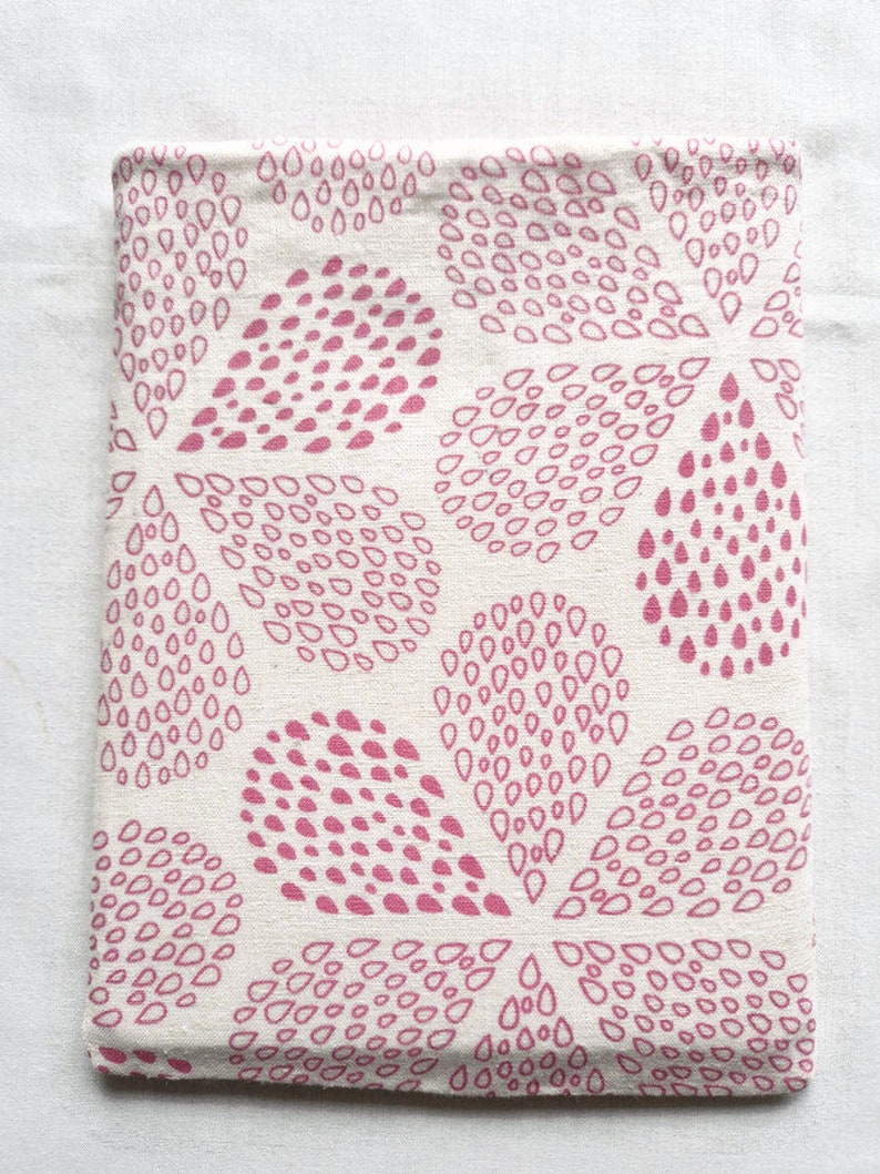 Puede incluir: Una funda rectangular de tela con una base de color blanco roto y un patr&oacute;n repetitivo de formas de gotas de color rosa. Las gotas est&aacute;n dispuestas en grupos que forman motivos m&aacute;s grandes similares a p&eacute;talos sobre el tejido.