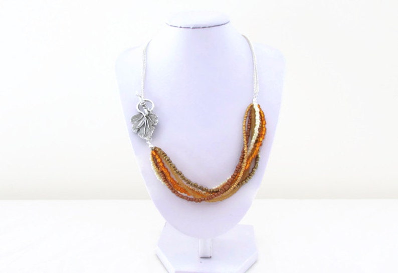 Op de afbeelding: Een meerstrengs ketting met bruine, oranje en cr&egrave;me kralen. De ketting heeft een zilveren bladeren hanger.