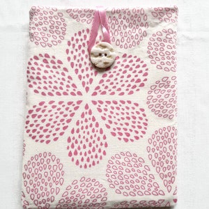 Puede incluir: Una funda de tela rectangular con un patr&oacute;n repetitivo de gotas rosas sobre un fondo blanco roto. La funda se asegura con un lazo de cinta rosa claro y un bot&oacute;n circular texturizado de color blanco roto centrado en el borde superior.