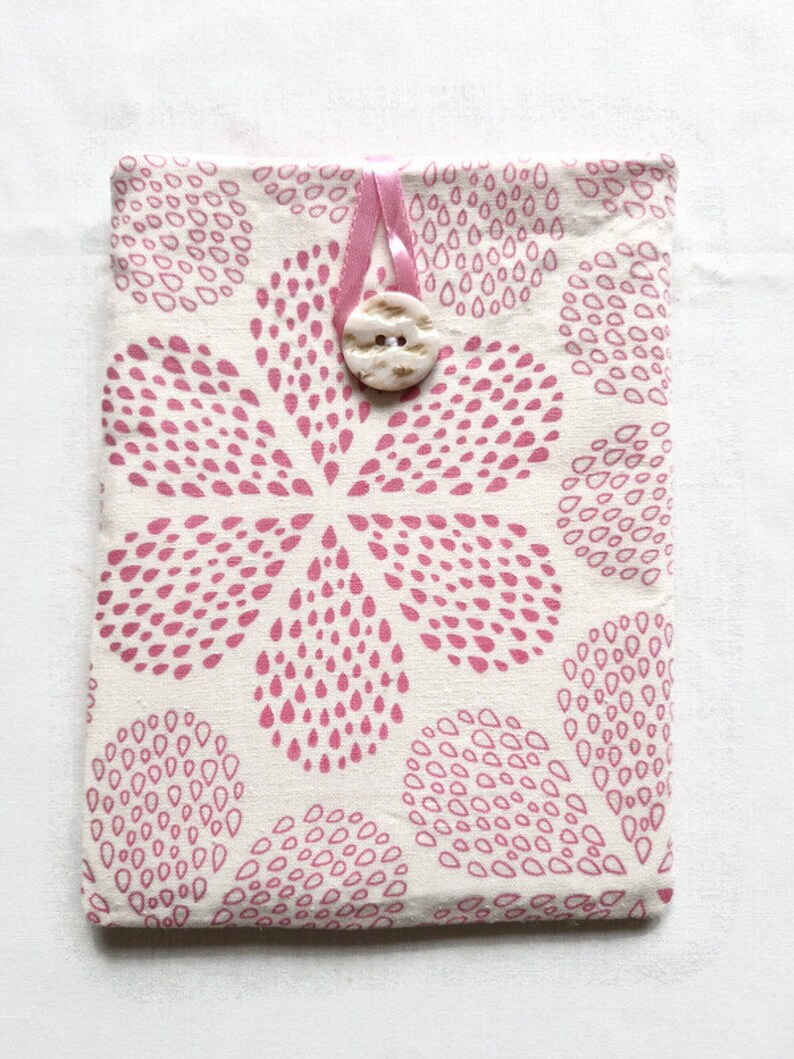 Puede incluir: Una funda de tela rectangular con un patr&oacute;n repetitivo de gotas rosas sobre un fondo blanco roto. La funda se asegura con un lazo de cinta rosa claro y un bot&oacute;n circular texturizado de color blanco roto centrado en el borde superior.