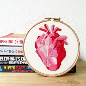 May include: A pink and red geometric heart embroidered on white fabric in a wooden embroidery hoop. The hoop is sitting on a stack of books.