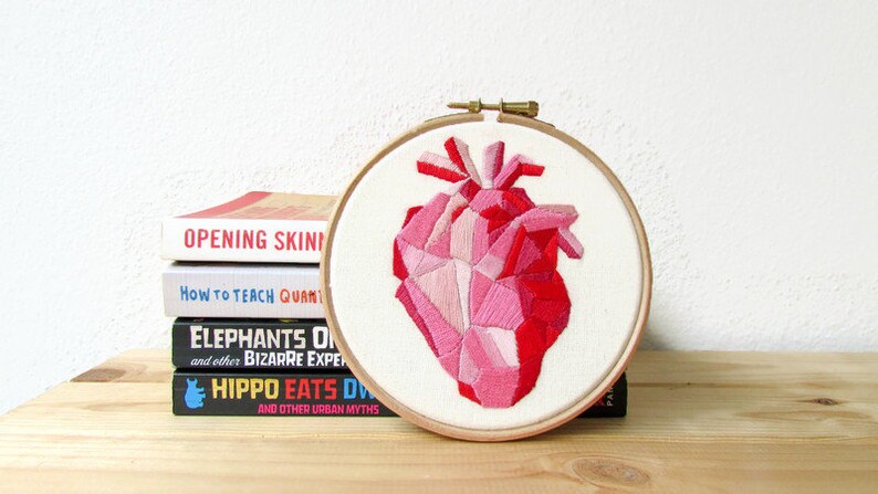 May include: A pink and red geometric heart embroidered on white fabric in a wooden embroidery hoop. The hoop is sitting on a stack of books.