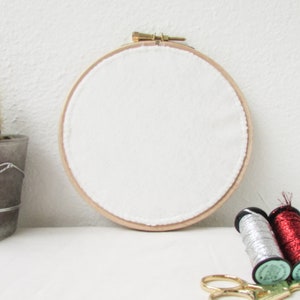 May include: A white embroidery hoop with a wooden frame, two spools of thread, one silver and one red, and a pair of gold scissors on a white surface.