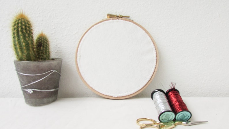 May include: A white embroidery hoop with a wooden frame, two spools of thread, one silver and one red, and a pair of gold scissors on a white surface.