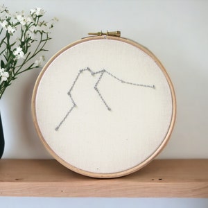 May include: A white embroidery hoop with a silver thread design of the Aquarius constellation. The hoop is on a wooden shelf with a vase of white flowers.