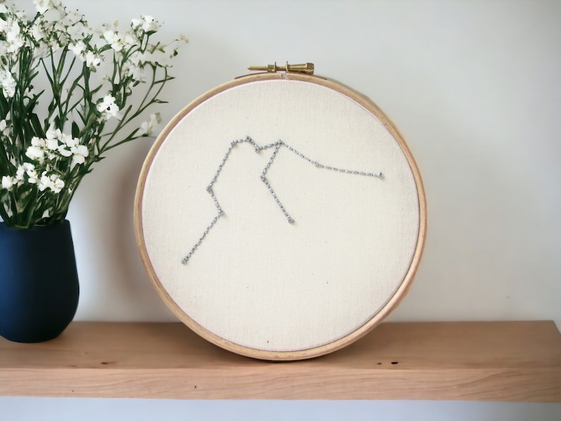 May include: A white embroidery hoop with a silver thread design of the Aquarius constellation. The hoop is on a wooden shelf with a vase of white flowers.