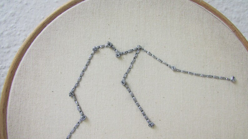 May include: A silver glitter embroidery of the Aquarius constellation on a white fabric background.