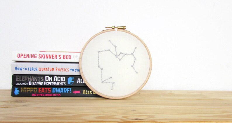 May include: A white embroidery hoop with a beige fabric background featuring a silver embroidered constellation of Sagittarius. The hoop is sitting on a wooden surface in front of a stack of four books.