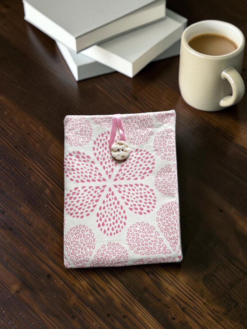 Puede incluir: Una funda de tela rectangular para libro con fondo blanco y un patr&oacute;n floral rosa repetitivo formado por peque&ntilde;as formas de gota. La funda se asegura con un lazo de cinta rosa que se sujeta sobre un bot&oacute;n blanco redondo y texturizado.