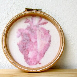 May include: A pink heart-shaped embroidery in a wooden embroidery hoop. The heart is a light pink colour and has a textured, watercolour-like appearance.