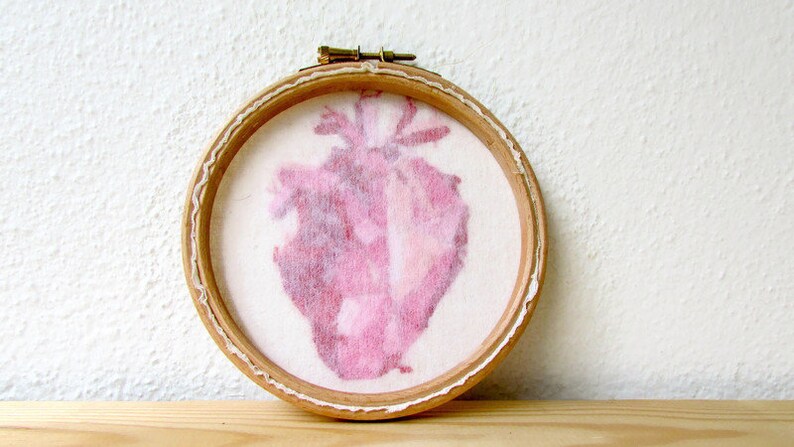 May include: A pink heart-shaped embroidery in a wooden embroidery hoop. The heart is a light pink colour and has a textured, watercolour-like appearance.