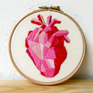 May include: A geometric heart embroidered on a white canvas in shades of pink and red. The heart is framed in a wooden hoop.