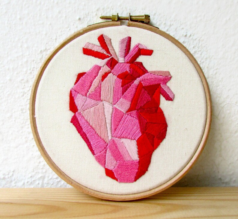 May include: A geometric heart embroidered on a white canvas in shades of pink and red. The heart is framed in a wooden hoop.