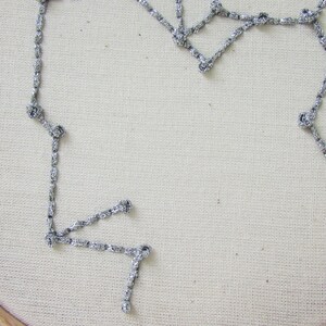 May include: A silver chain stitched onto a white fabric background in the shape of a constellation.