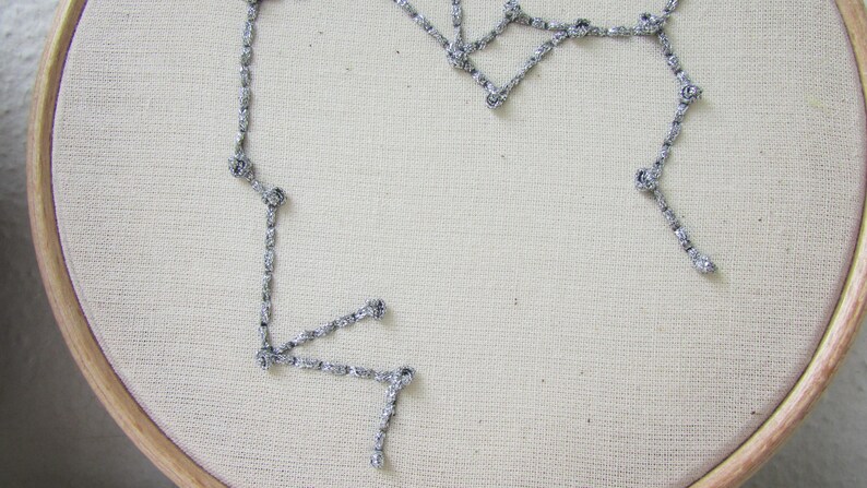 May include: A silver chain stitched onto a white fabric background in the shape of a constellation.