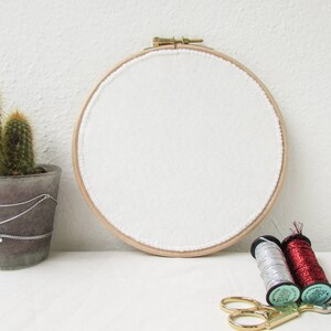 May include: A white embroidery hoop with a wooden frame and white fabric stretched across it. There are two spools of thread, one silver and one red, and a pair of gold scissors on a white surface.