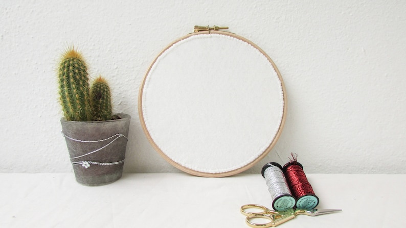 May include: A white embroidery hoop with a wooden frame and white fabric stretched across it. There are two spools of thread, one silver and one red, and a pair of gold scissors on a white surface.