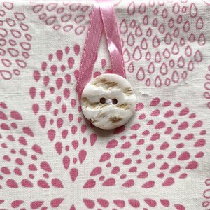 Puede incluir: Una bolsa de tela color crema con un patr&oacute;n repetitivo de formas de gota rosas dispuestas en racimos florales. Un lazo de cinta de sat&eacute;n rosa est&aacute; asegurado por un bot&oacute;n redondo de color blanco roto con marcas marrones y dos agujeros.