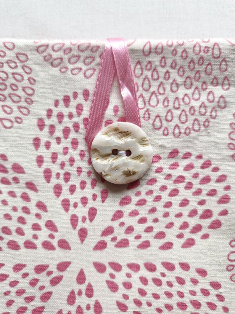 Puede incluir: Una bolsa de tela color crema con un patr&oacute;n repetitivo de formas de gota rosas dispuestas en racimos florales. Un lazo de cinta de sat&eacute;n rosa est&aacute; asegurado por un bot&oacute;n redondo de color blanco roto con marcas marrones y dos agujeros.