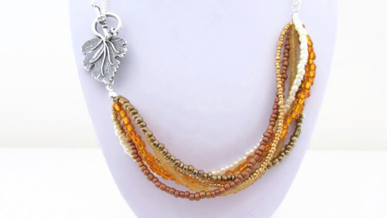 Op de afbeelding: Een meerstrengs ketting met een zilveren bladeren hanger. De ketting is gemaakt van kleine kralen in tinten goud, bruin en wit.