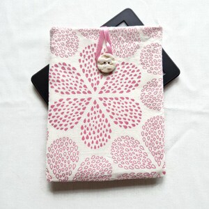 Puede incluir: Una funda de tela rectangular con fondo crema y un patr&oacute;n repetitivo de gotas rosas que forman una gran flor. La funda se asegura con un lazo de cinta rosa y un bot&oacute;n redondo de color blanco roto con una superficie texturizada.