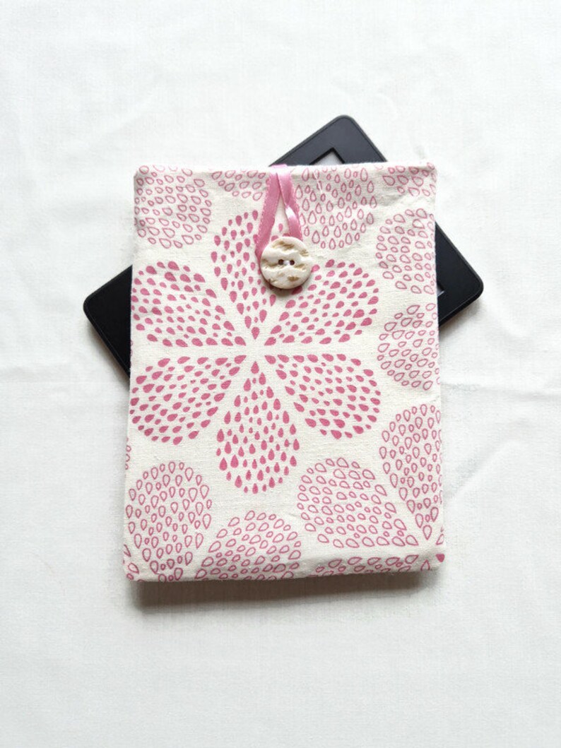 Puede incluir: Una funda de tela rectangular con fondo crema y un patr&oacute;n repetitivo de gotas rosas que forman una gran flor. La funda se asegura con un lazo de cinta rosa y un bot&oacute;n redondo de color blanco roto con una superficie texturizada.