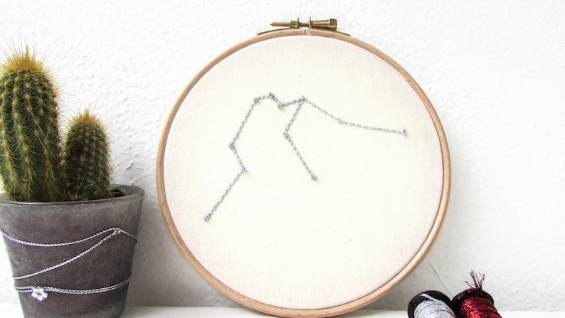 May include: A white embroidery hoop with a silver thread design of the Aquarius constellation. The hoop is hanging on a white wall.
