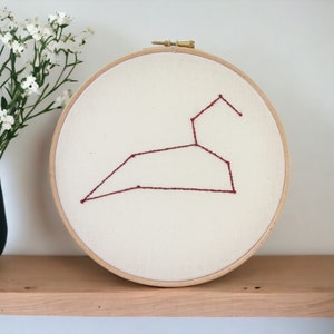 May include: A round embroidery hoop featuring the Leo constellation stitched in burgundy thread on a cream-colored fabric. The wooden hoop is displayed on a wooden shelf, with a blue vase of white flowers to the left.