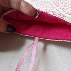 Puede incluir: Una bolsa de tela con un exterior de color crema con un patr&oacute;n repetitivo de puntos rosas y un forro interior de color rosa intenso. La bolsa incluye un lazo de cinta rosa claro unido al borde inferior para colgarla o transportarla.