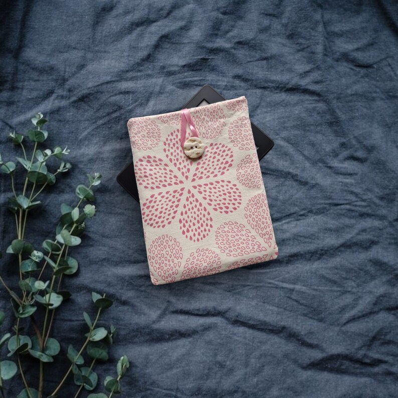 Puede incluir: Una funda de tela rectangular con fondo crema y un patr&oacute;n floral rosa repetitivo hecho de peque&ntilde;os puntos. La funda se asegura con un lazo de cinta rosa y un bot&oacute;n texturizado de color claro en la parte superior central.