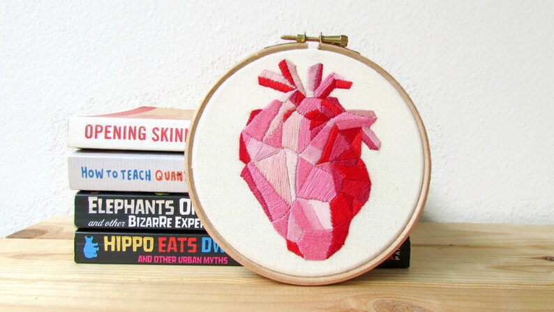 May include: A pink and red geometric heart embroidered on white fabric in a wooden embroidery hoop. The hoop is sitting on a stack of books.
