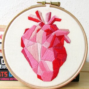 May include: A geometric heart embroidered in shades of pink and red on white fabric, framed in a wooden hoop.