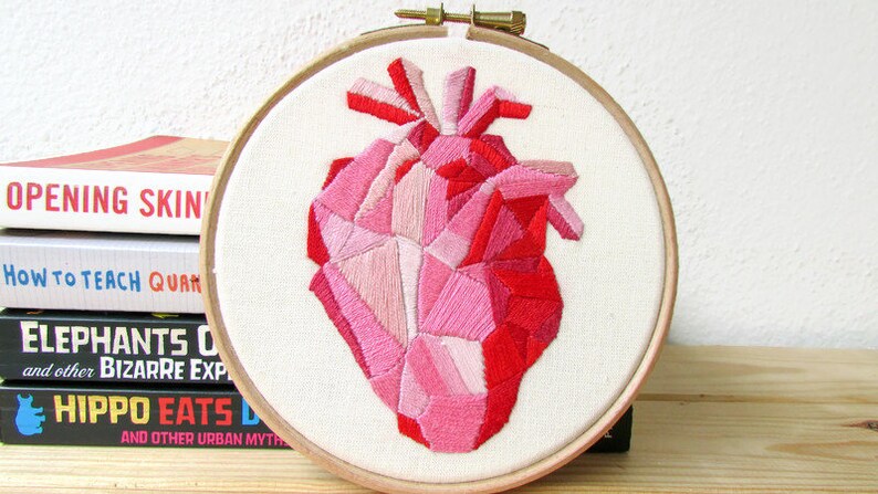 May include: A geometric heart embroidered in shades of pink and red on white fabric, framed in a wooden hoop.