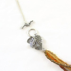 Op de afbeelding: Een meerstrengs ketting met een zilveren bladeren hanger en een zilveren schuifgesp. De ketting is gemaakt van kleine, ronde kralen in tinten bruin, beige en goud.