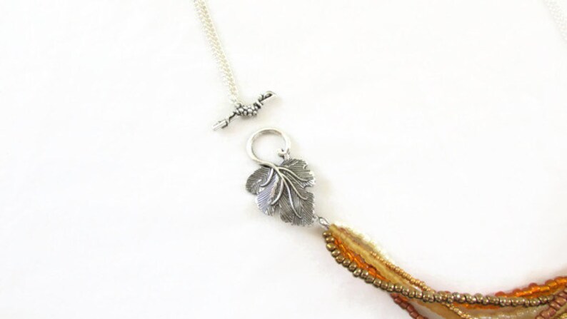 Op de afbeelding: Een meerstrengs ketting met een zilveren bladeren hanger en een zilveren schuifgesp. De ketting is gemaakt van kleine, ronde kralen in tinten bruin, beige en goud.