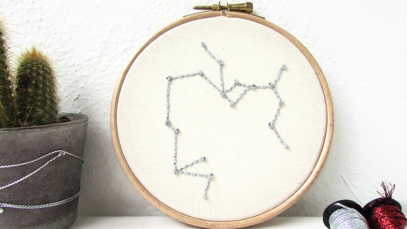 May include: A white embroidery hoop with a silver thread design of the Sagittarius constellation. The hoop is on a white surface with a potted cactus and spools of thread in the background.