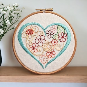 May include: A teal heart-shaped embroidery hoop with a floral design in shades of orange, yellow, red, and green. The hoop is on a wooden shelf.