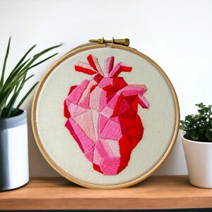 May include: A pink and red geometric heart embroidered on a white fabric stretched over a wooden embroidery hoop.