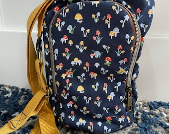 Op de afbeelding: Een klein blauw canvas tasje met een gele schouderband en een print van paddenstoelen. De tas heeft een ritssluiting en een klein vakje aan de voorkant.