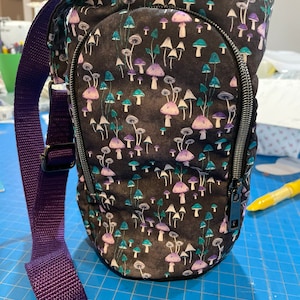 Peut inclure: Un sac en tissu noir avec une bandoulière violette et une fermeture éclair. Le sac a un imprimé de champignons avec des champignons violets, verts et blancs sur un fond noir.