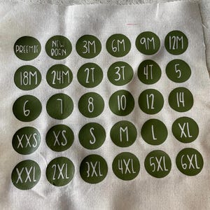 Può includere: Un campione di tessuto beige chiaro con cerchi verdi che mostrano taglie in testo bianco. Le taglie vanno da "PREEMIE" a "6XL", e includono taglie numeriche da 5 a 14. Il testo include anche "NEWBORN", "3M", "6M", "9M", "12M", "18M", "24M", "2T", "3T" e "4T".